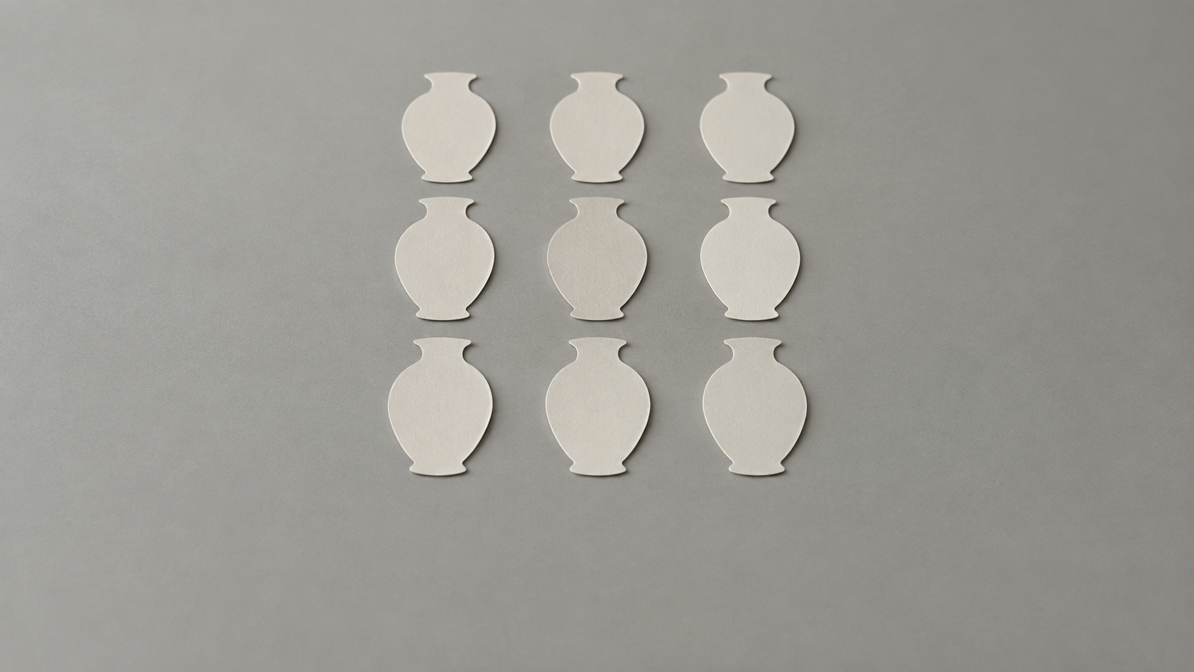 Nine identical plain paper cutout shapes of one unlabelled vase arranged in a neat three-by-three grid on a neutral grey studio surface